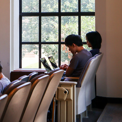 Two students work on laptops at a table beside large windows overlooking trees on the Rice University campus.
