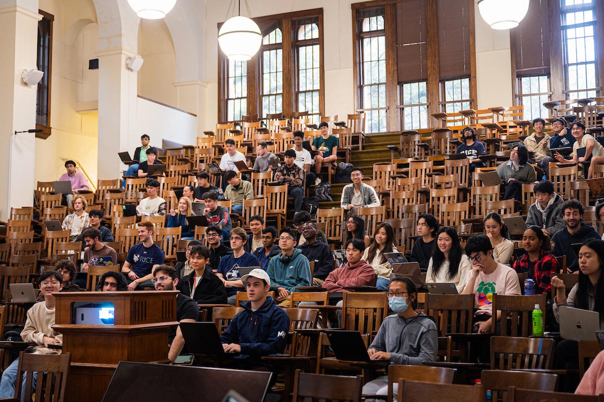 A lecture hall full of college students sits in rows of wooden desks.